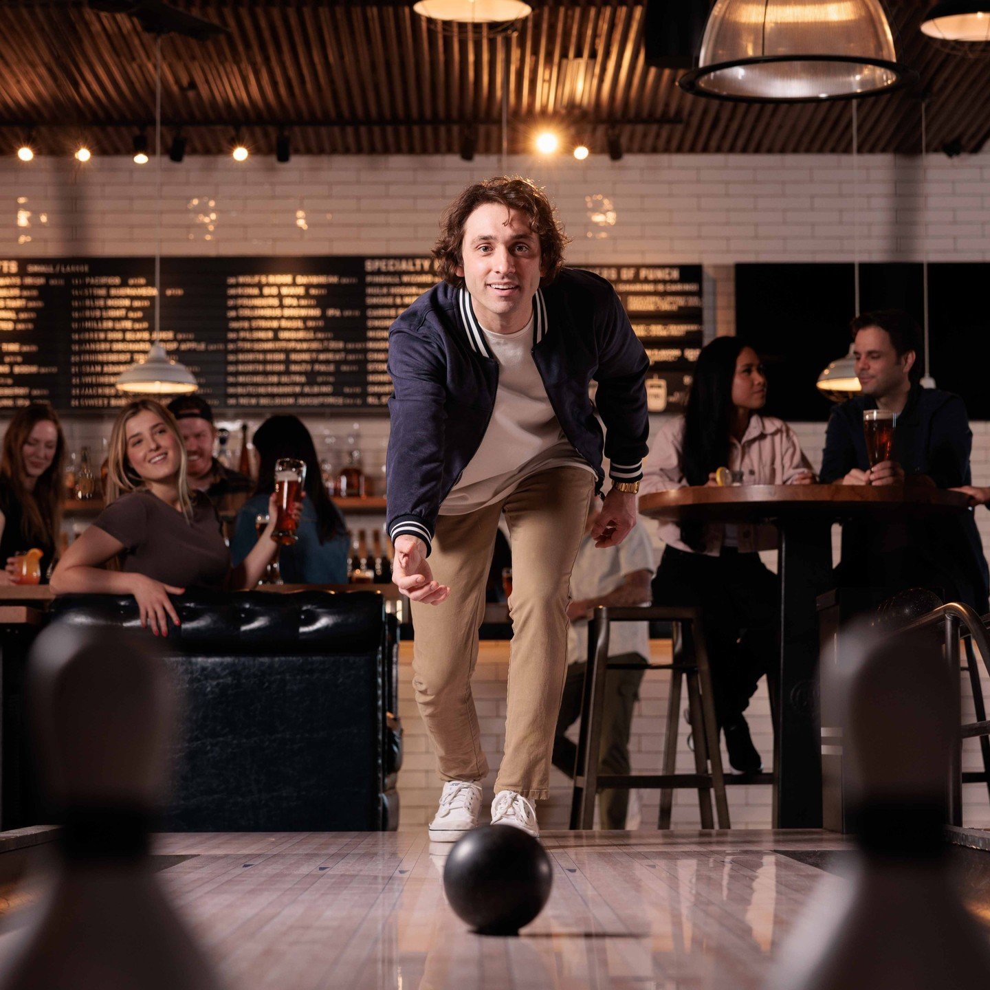 Bowler rolling on a wooden lane while friends watch from tables inside Pins.