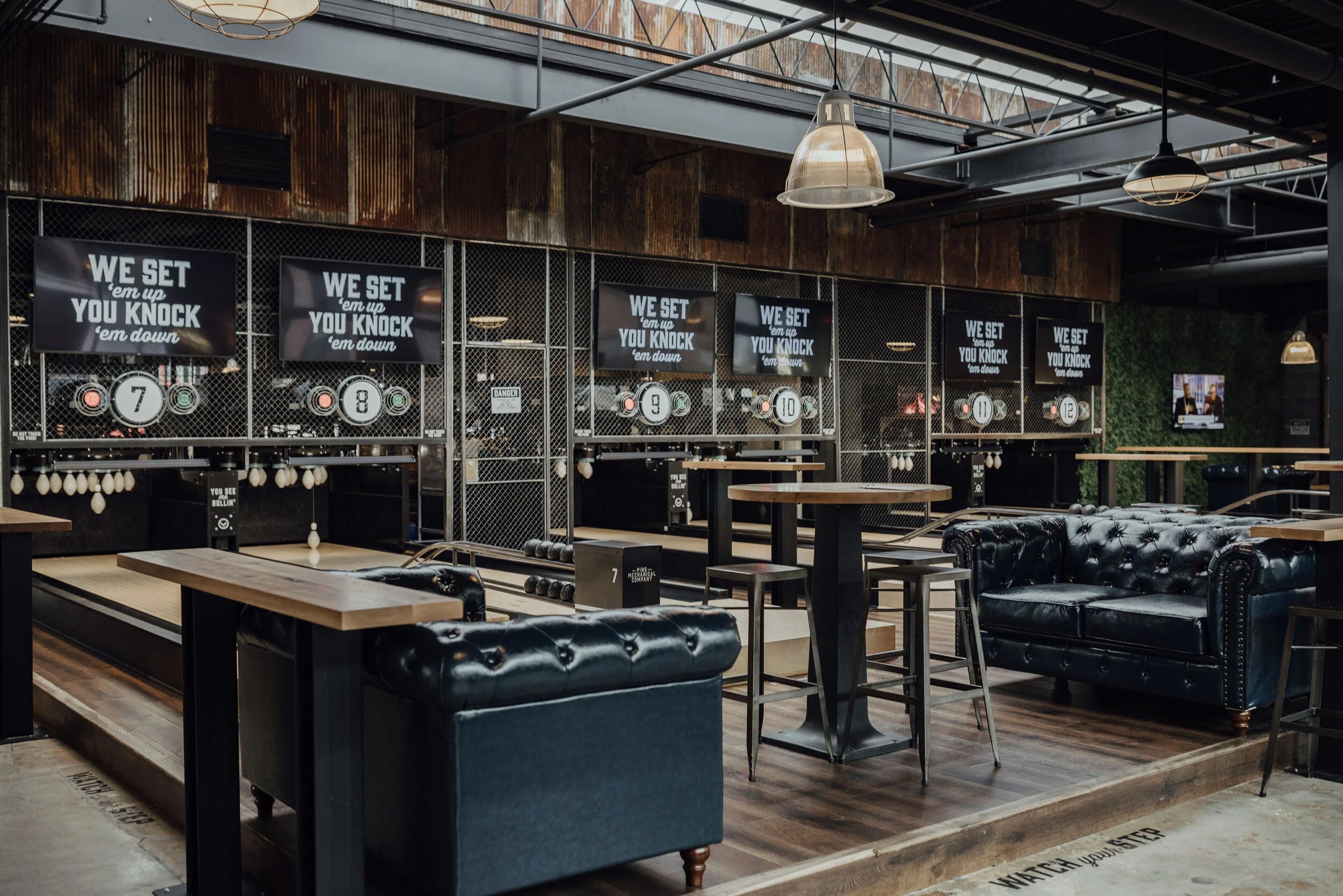 Pins bowling interior with leather sofas and score screens reading “We set ’em up, you knock ’em down.”