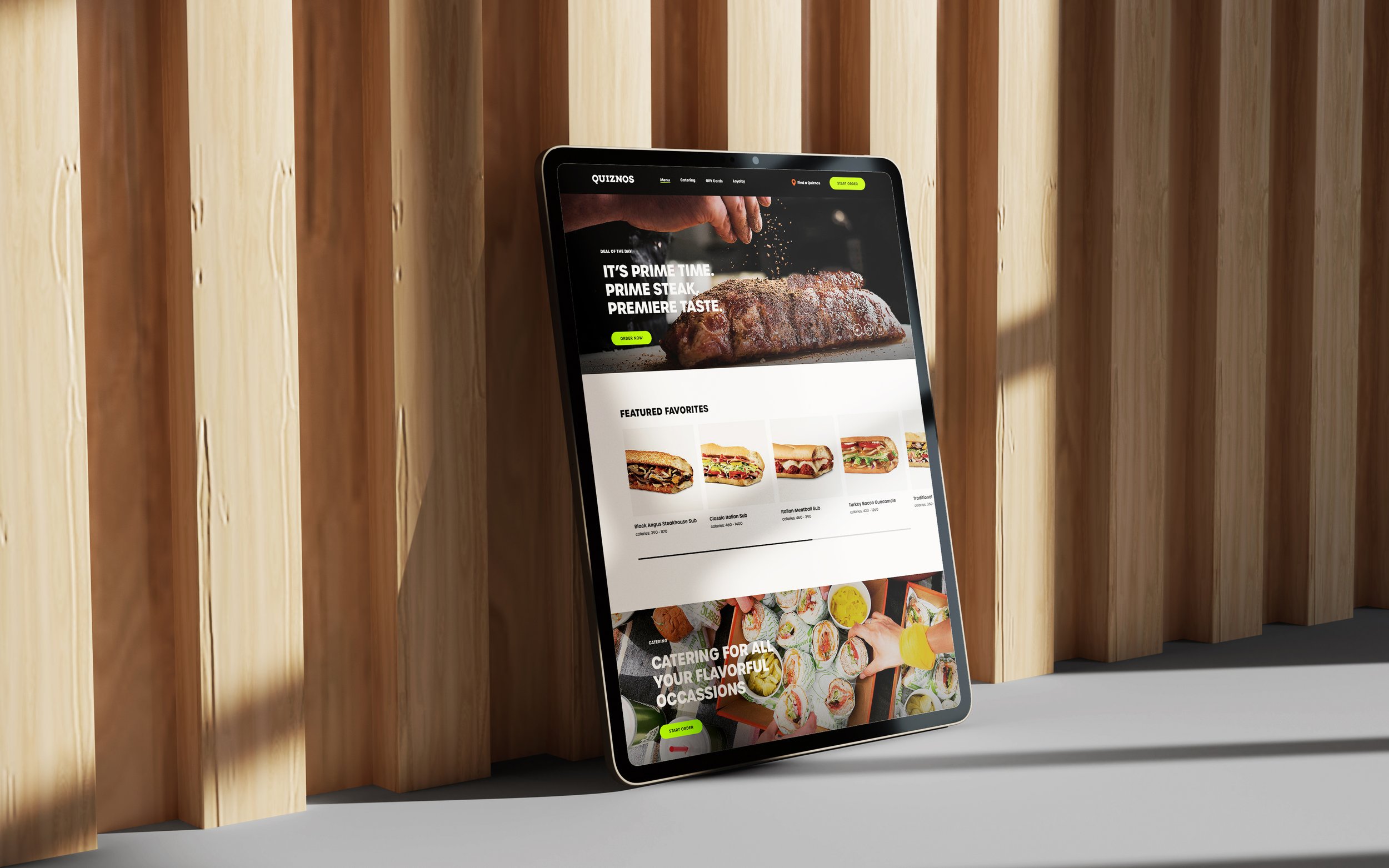 Tablet leaning against a wooden wall, showing the Quiznos site with sandwiches and a steak.