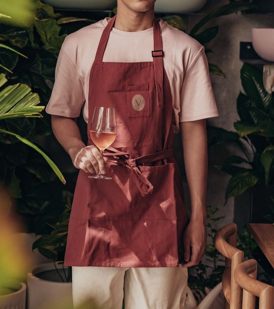 Person in a light shirt and red apron holding a glass of rosé.